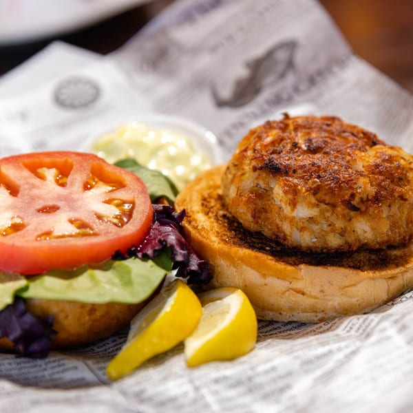 A crab cake sandwich on a bun with lettuce, tomato, lemon wedges, and tartar sauce.