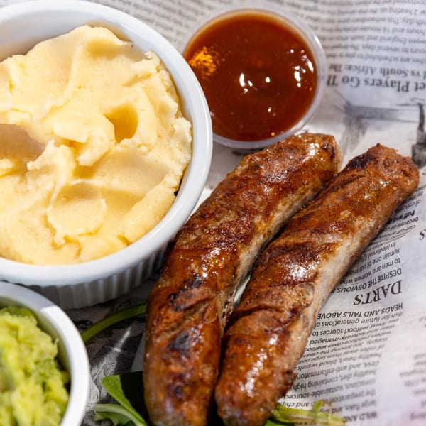 Two grilled sausages, mashed potatoes, mushy peas, and brown sauce on newspaper.