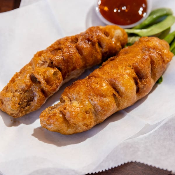 Two golden-brown battered sausages on white paper with a side of red sauce and greens.