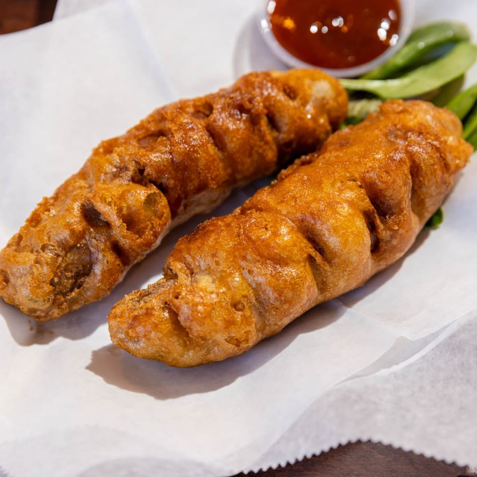 Two golden-brown battered sausages on white paper with a side of red sauce and greens.