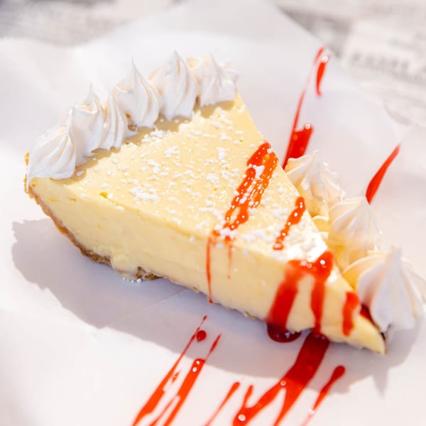 Slice of key lime pie with whipped cream, powdered sugar, and raspberry drizzle on white paper.