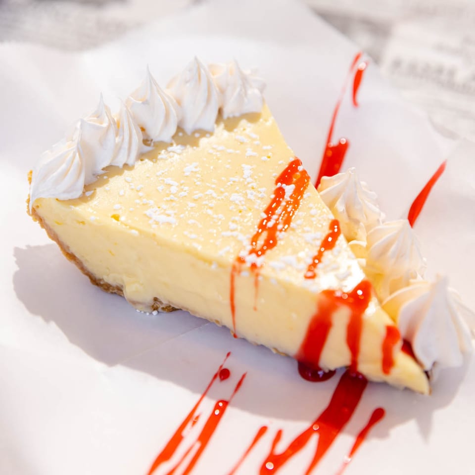 Slice of key lime pie with whipped cream, powdered sugar, and raspberry drizzle on white paper.
