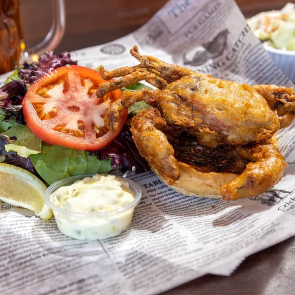 A fried soft-shell crab sandwich with lettuce, tomato, lemon, and tartar sauce on newspaper.