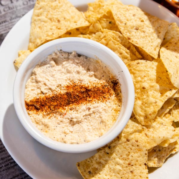 A white bowl of hummus with paprika and black pepper, surrounded by tortilla chips.
